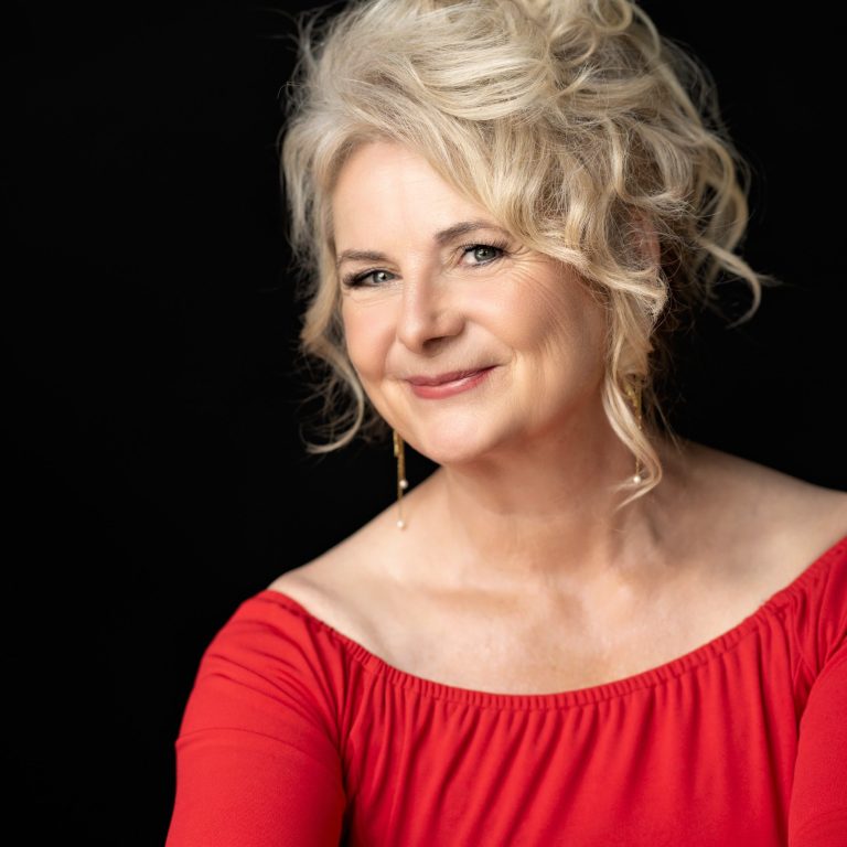 Constanze Yacine Frau mit blonden Locken, rotem Oberteil und freundlichem Lächeln, vor schwarzem Hintergrund.