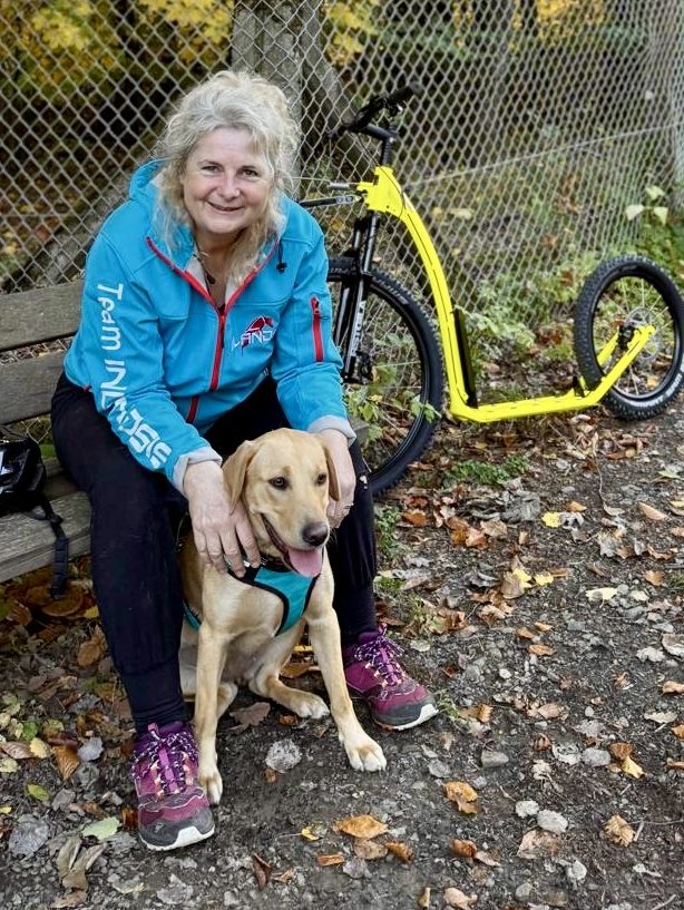 Cookie und Constanze beim Dogscooting Eine Frau mit blonem Haar sitzt lächelnd auf einer Bank und streichelt ihren Labrador neben einem gelben Dogscooter.